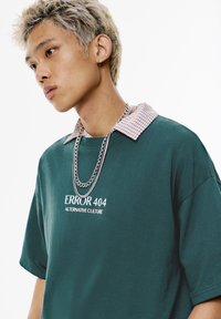 Green oversized t-shirt with a checkered collar, featuring the text "ERROR 404 ALTERNATIVE CULTURE" and layered silver chain necklace.