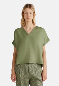 Top verde in lino a V con maniche corte arrotolate, abbinato a pantaloni verdi a fantasia con un design a rombi in verde scuro.