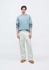 Ανοιχτό μπλε φούτερ με V-λαιμόκοψη και μανίκια ράγκλαν, συνδυασμένο με ανοιχτό γκρι παντελόνι φαρδιάς γραμμής και ταμπά ξύλινα σανδάλια· ομοιόμορφη υφή σε όλο το ρούχο.