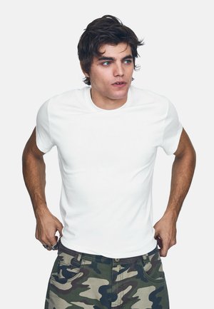 Jeune homme aux cheveux foncés portant un t-shirt blanc et un pantalon camouflage, ajustant son pantalon et regardant sur le côté sur un fond uni.