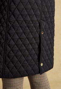 Manteau matelassé noir avec couture en losanges et boutons-pression dorés sur un pantalon à motif pied-de-poule sur fond beige.
