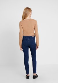 Mujer de pie de espaldas, vistiendo una blusa de manga larga color beige, jeans skinny azules oscuros y mocasines negros, frente a un fondo blanco liso.