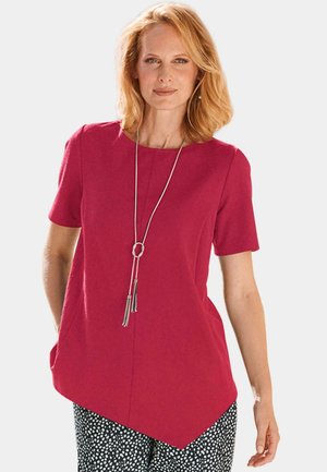 Haut fuchsia à manches courtes et décolleté arrondi. Il présente un ourlet asymétrique et un long collier en argent avec des franges.