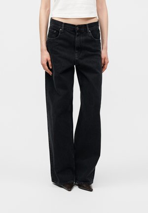 Osoba nosiaca čierne džínsy s vysokým pásom a širokými nohavicami s prednými vreckami, v kombinácii s tmavými špicatými topánkami a krátkym bielym topom.