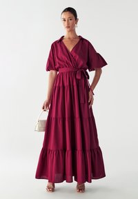 Burgundská maxi šaty s krátkými nabíranými rukávy, uvázaným pasem a vrstvenými volány na spodním lemu. Hladká tkanina s lehkou strukturou.