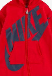 Crvena zip-up dukserica izrađena od mekanog materijala, sa velikim tamnoplavi Nike logom na prednjoj strani i kengur džepom na struku.