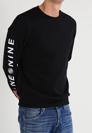 Sweatshirt noir avec un col rond et des poignets côtelés, arborant une inscription blanche audacieuse sur la manche gauche et une coupe décontractée. Porté avec un jean bleu.