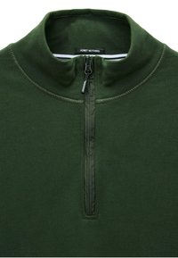 Ciemnozielony sweter z wysokim kołnierzem, półzamkiem i miękkim, teksturowanym materiałem. Posiada metkę przy szyi z napisem "NIE PRZYZNAJ SIĘ DO NICZEGO."