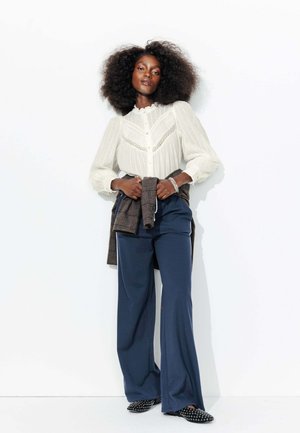 Chemisier blanc texturé avec des accents en dentelle, associé à un pantalon large bleu marine. Des chaussures noires à clous complètent la tenue, accompagnée d'une veste en plaid nouée.