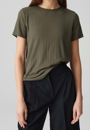 Femme portant un haut côtelé à manches courtes vert olive, légèrement rentré dans un pantalon noir plissé à taille haute, debout devant un fond uni.