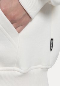 Hvid hætte sweatshirt med ribbet manchet, en frontlomme og et lille sort label med "smilodox" på sidesømmen. Struktureret stof.