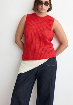 Kobieta ubrana w czerwoną, bezrękawną dzianinową bluzkę na kremowej koszuli i ciemnych, szerokich dżinsach, z krótkimi włosami i owalnymi okularami przeciwsłonecznymi, stojąca na tle białej ściany.