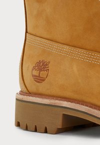 Gros plan sur le talon d'une botte en daim beige avec une semelle en caoutchouc brun épaisse et un logo d'arbre embossé près d'une double couture blanche.