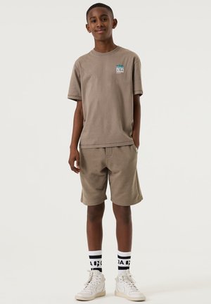 Ragazzo in piedi che sorride, indossa una maglietta marrone con logo a palma, pantaloncini abbinati, calzini bianchi con testo nero e scarpe da ginnastica bianche.