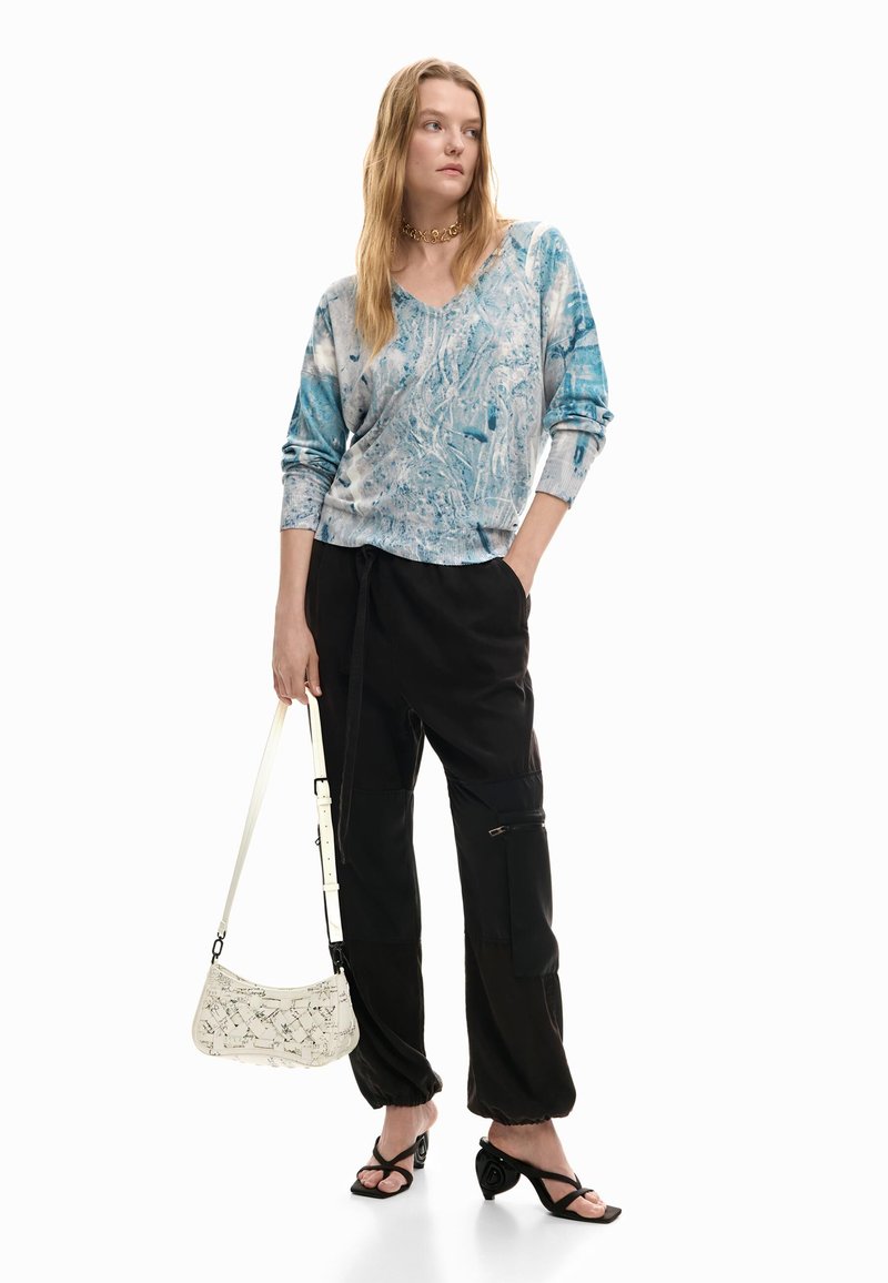 Mujer con una blusa de manga larga azul y blanca con efecto mármol, pantalones cargo negros, sandalias negras de tacón, collar de cadena dorada y un bolso blanco con patrones.