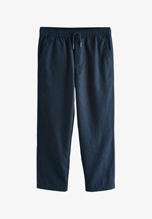 Barn iført marineblå bukser, hvide sneakers og en hvid t-shirt med den ene hånd i lommen mod en ensfarvet beige baggrund.