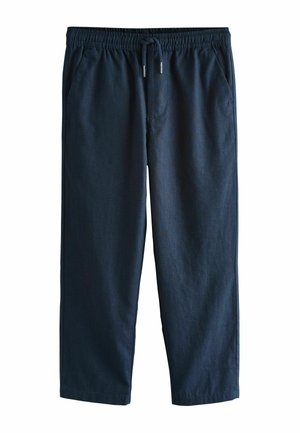 Barn iført marineblå bukser, hvide sneakers og en hvid t-shirt med den ene hånd i lommen mod en ensfarvet beige baggrund.