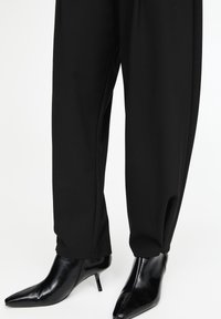 Bottines noires à bout pointu avec talons kitten, portées avec un pantalon noir droit sur fond blanc.