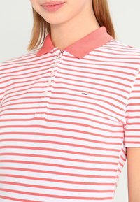 Růžový a bílý proužkovaný polo tričko s zapínacím límcem, obsahující malé logo na hrudi a texturovanou látku.