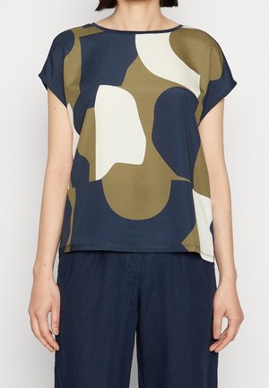 Femme portant un haut ample à manches courtes avec un motif abstrait bleu marine, olive et blanc, associé à un pantalon bleu foncé, debout sur un fond uni.