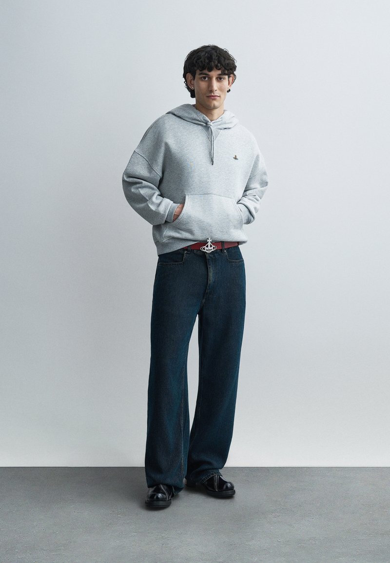 Sweat à capuche gris avec une poche frontale et un cordon, associé à un jean denim foncé. La tenue est complétée par une ceinture rouge à boucle métallique.