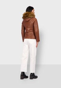 Bruine gewatteerde jas met een nep-bont capuchon, gecombineerd met witte wijdere broeken en zwarte chunky haklaarzen. Achteraanzicht weergegeven.