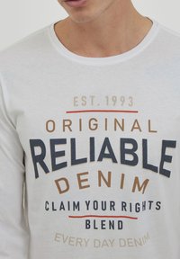 Hvit langermet T-skjorte med rund hals og fet svart, brun og rød tekst som sier "PÅLITELIG DENIM" og "GRUNNLAGT 1993."