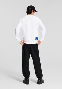 Weißes Sweatshirt mit gerippten Bündchen und Saum, mit einem blauen Etikett am unteren Rücken. Kombiniert mit schwarzen, schmal zulaufenden Jogginghosen und schwarzen Stiefeln.