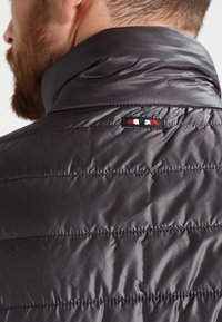 Dunkelgraue Steppjacke mit genoppter Textur, hohem Kragen und einem kleinen gestickten Logo mit roten, weißen und blauen Akzenten auf dem Rücken.
