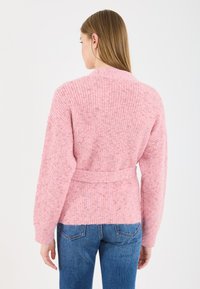 Rožā adīta džemperis ar jostu viduklī, ar ribotām tekstūrām un nokristām plecu līnijām, apvienots ar ziliem džinsiem.