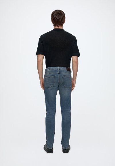 Homme debout de dos, portant une chemise noire à manches courtes, un jean bleu avec des poches arrière et des chaussures noires sur un fond blanc uni.