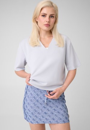 Femme blonde portant un haut gris clair à manches courtes et une mini-jupe bleue à motifs, debout devant un fond gris uni.