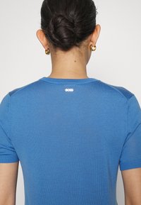 Mujer con cabello oscuro recogido en un moño, usando pendientes de aro dorados y una camiseta de manga corta azul con el logo "BOSS" en la parte superior de la espalda.
