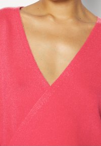 Haut en tricot rose à col en V, avec une texture douce et un design croisé ; le tissu présente un léger éclat pour plus de détails.