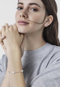 Bracciale d'argento con accenti floreali e perle indossato su un polso, abbinato a orecchini a forma di fiore. Modella con felpa grigio chiaro.