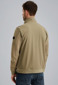 Giacca da uomo color khaki con zip e colletto alto, e polsini elasticizzati. Texture liscia con una toppa nera con logo sulla manica sinistra.