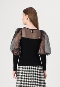 Zwarte fitted top met doorzichtige puff mouwen, versierd met witte parels. De achterkant heeft een sleutelgaatje en wordt gecombineerd met een houndstooth rok.