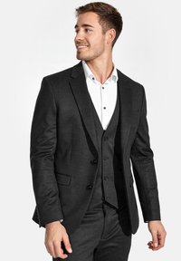 Maßgeschneiderter dunkelgrauer Anzug aus glattem, strukturiertem Stoff. Dreiknopf-Weste unter einem taillierten Blazer, ergänzt durch ein weißes Hemd und braune Knopfakzente.