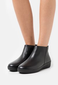 Botas de tobillo de cuero negro con textura suave, tacón bajo de cuña y puntera redondeada. Diseño sin costuras con ningún hardware o patrones visibles.