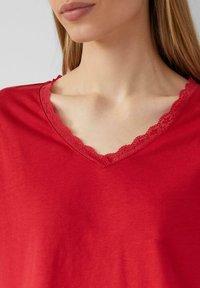 Haut rouge à col en V avec une bordure en dentelle au niveau de l'encolure, fabriqué dans un tissu doux à la texture lisse, offrant un ajustement décontracté et confortable.