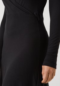 Svart klänning med långa ärmar, omlottmodell och knytning i midjan. Tillverkad av mjukt, stretchigt material med en slät textur.