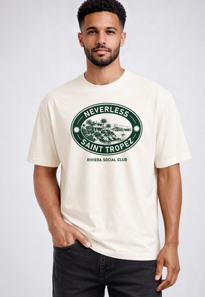 Mann mit kurzen lockigen Haaren und Bart, der ein weißes T-Shirt mit grünem "Nevertheless Saint Tropez Riviera Social Club" Text und Küstengrafik trägt.