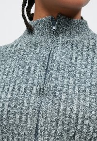 Pelēks teksturēts augšdaļa ar rāvējslēdzēju un augstu apkakli, ar kontrastējošu gaišāku rakstu un melnas krāsas rāvējslēdzēja detaļu.