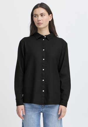 Jonge vrouw in een zwarte knoopsluitende blouse met lange mouwen en lichtblauwe jeans, die iets naar links kijkt tegen een effen achtergrond.