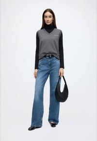 Šedý tílko přes černé tričko s dlouhým rukávem, doplněné světle modrými širokými džínami. Drží černou zakřivenou kabelku.
