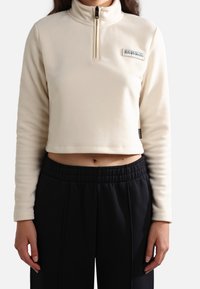 Sudadera corta de polar color crema con cremallera media, cuello alto y un pequeño parche de logo de la marca en el pecho. Tejido texturizado y suave.