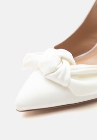 Λευκό μπαλαρίνα παπούτσι με μυτερή μύτη και μεγάλο, μαλακό φιόγκο από ύφασμα στο πάνω μέρος μπροστά.