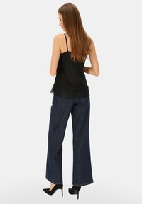Czarna jedwabna koszulka na ramiączkach z koronkową lamówką, zestawiona z szerokimi spodniami z ciemnego denimu i czarnymi szpilkami. Widok od tyłu.