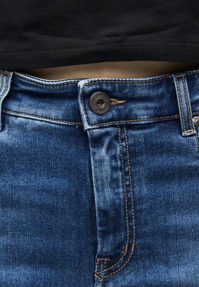 Gros plan sur le torse portant un jean bleu avec le bouton fermé et un haut noir légèrement relevé au-dessus de la taille.