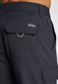 Närbild av svarta Columbia-shorts med en ficka med klaff och en metallöga på höften, bärs av en person med synlig midja och hud.